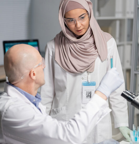 Young Muslim female virologist in hijab and whitecoat looking at blue liquid in flask held by mature male colleague in laboratory